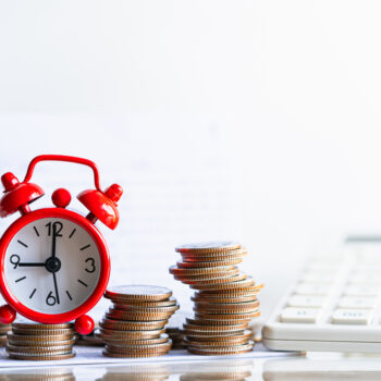 A small red alarm clock is placed among stacks of coins with a white calculator in the background, symbolizing the concepts of time, money, and financial planning.
