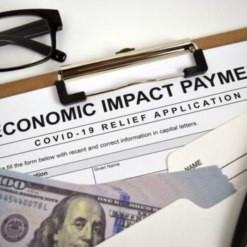 A clipboard with an Economic Impact Payment COVID-19 Relief Application form, a pair of eyeglasses, a pen, an envelope, and a stack of $100 bills.