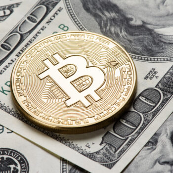 A gold-colored physical Bitcoin coin rests on top of several U.S. hundred-dollar bills, symbolizing the relationship between cryptocurrency and traditional currency.